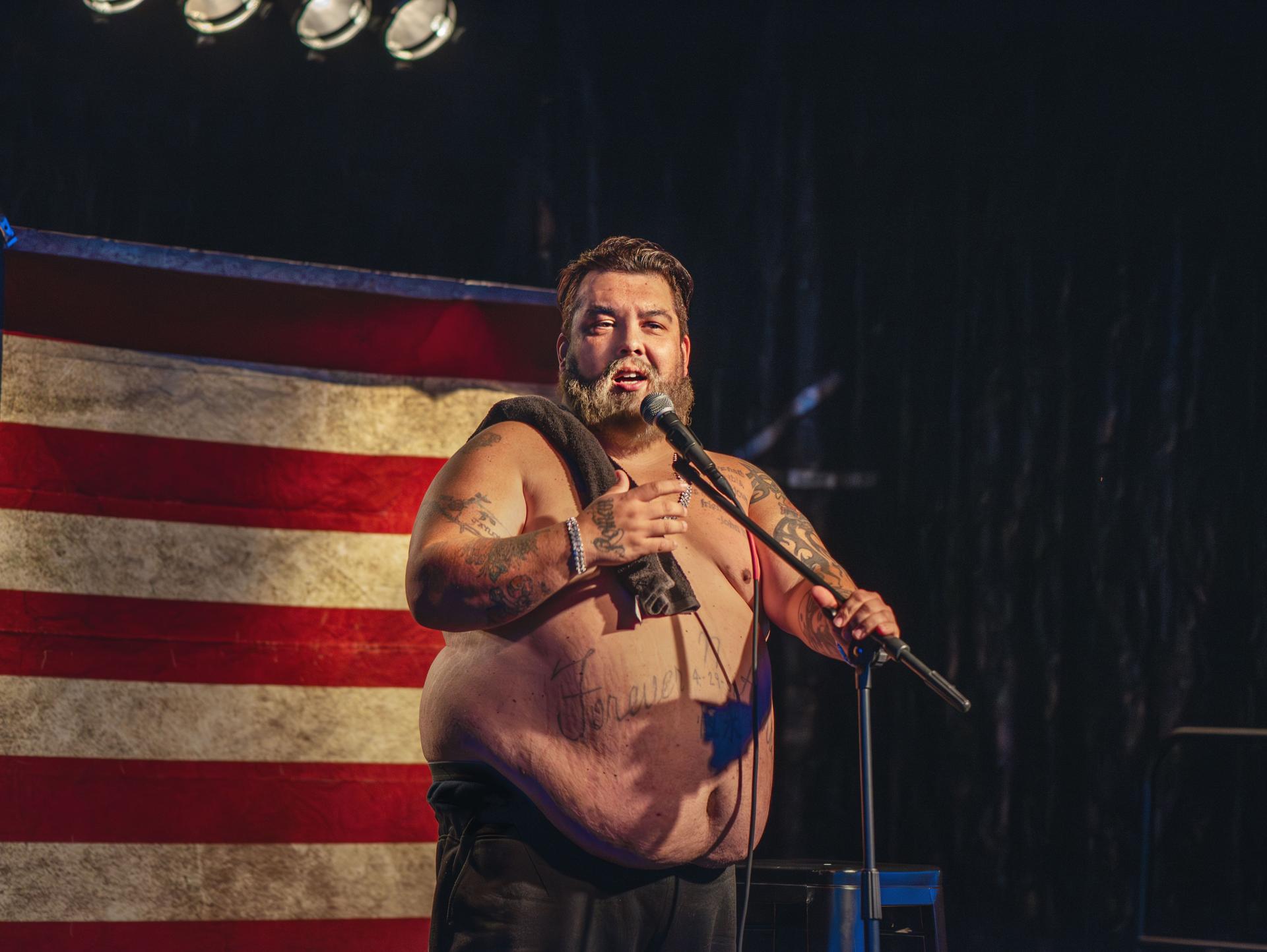 Stand-up comedian takes the stage shirtless, showcasing his tattoos as he entertains the crowd. A middle-aged performer confidently delivers his routine, with the American flag serving as a striking backdrop.